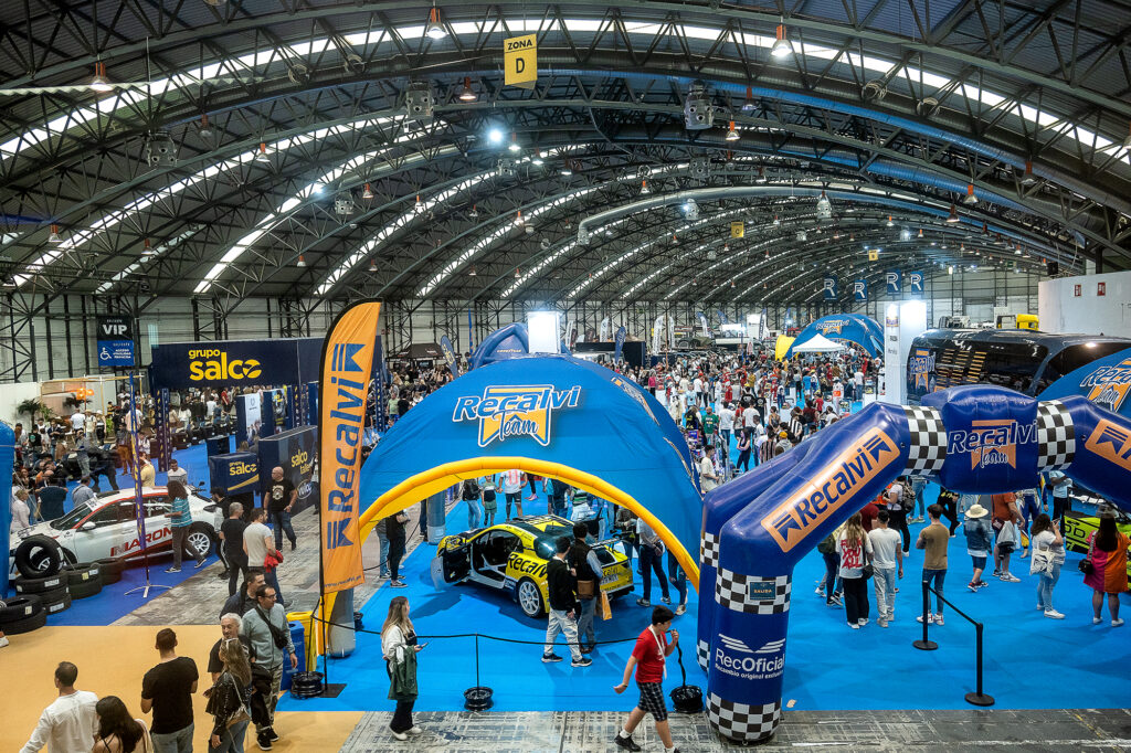 Busy indoor car exhibition with blue carpet, inflatable RecAlvi race cars, and numerous booths under a curved metal roof.