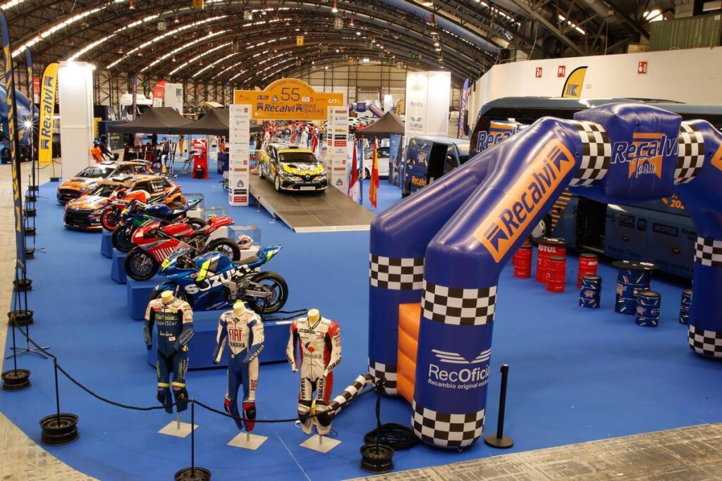 Indoor motorsport expo with rows of racing motorcycles on blue carpet and life-size racing-suit mannequins under a large blue Recalvi inflatable arch off to the side.